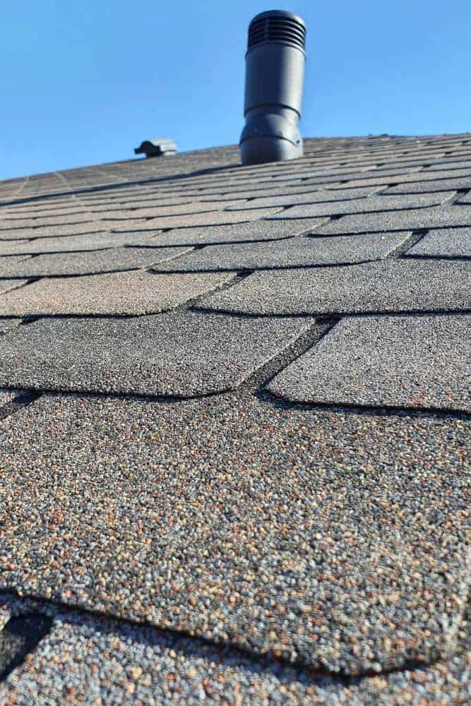 Close-up view of a sloped roof with asphalt shingles under a clear blue sky, expertly restored by leading restoration contractors. A black vent pipe protrudes near the roof's peak, with another recently refurbished roof structure visible in the background.