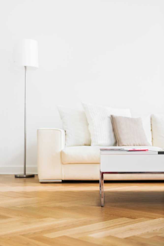 A modern living room features a white sofa with gray and beige cushions, a sleek white coffee table, and a tall, slender floor lamp. With light wood parquet flooring and a minimalist aesthetic, this space showcases the meticulous touch often seen from top restoration contractors.
