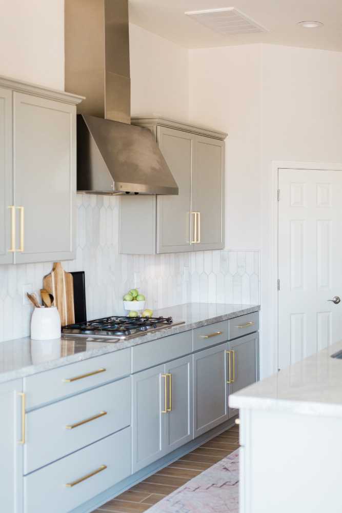Modern kitchen with light gray cabinets, gold handles, and a stainless steel range hood. A white backsplash features geometric tiles. The countertop holds a cutting board, apples, and a ceramic vase placed by restoration company experts after fixing previous water damage. A closed white door is in the background.