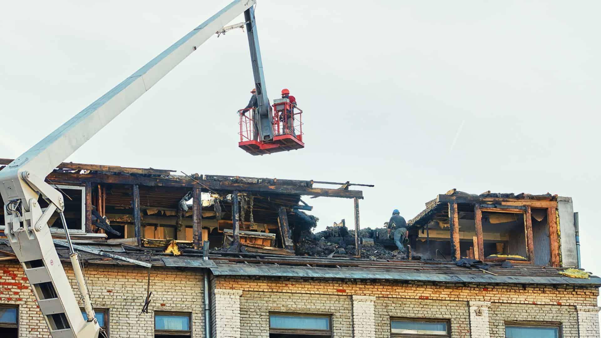 Construction workers are inspecting and repairing the damaged roof of a brick building. One worker is in a red lift, while others are on the roof surrounded by debris, indicating the building may have been affected by fire or storm damage.
