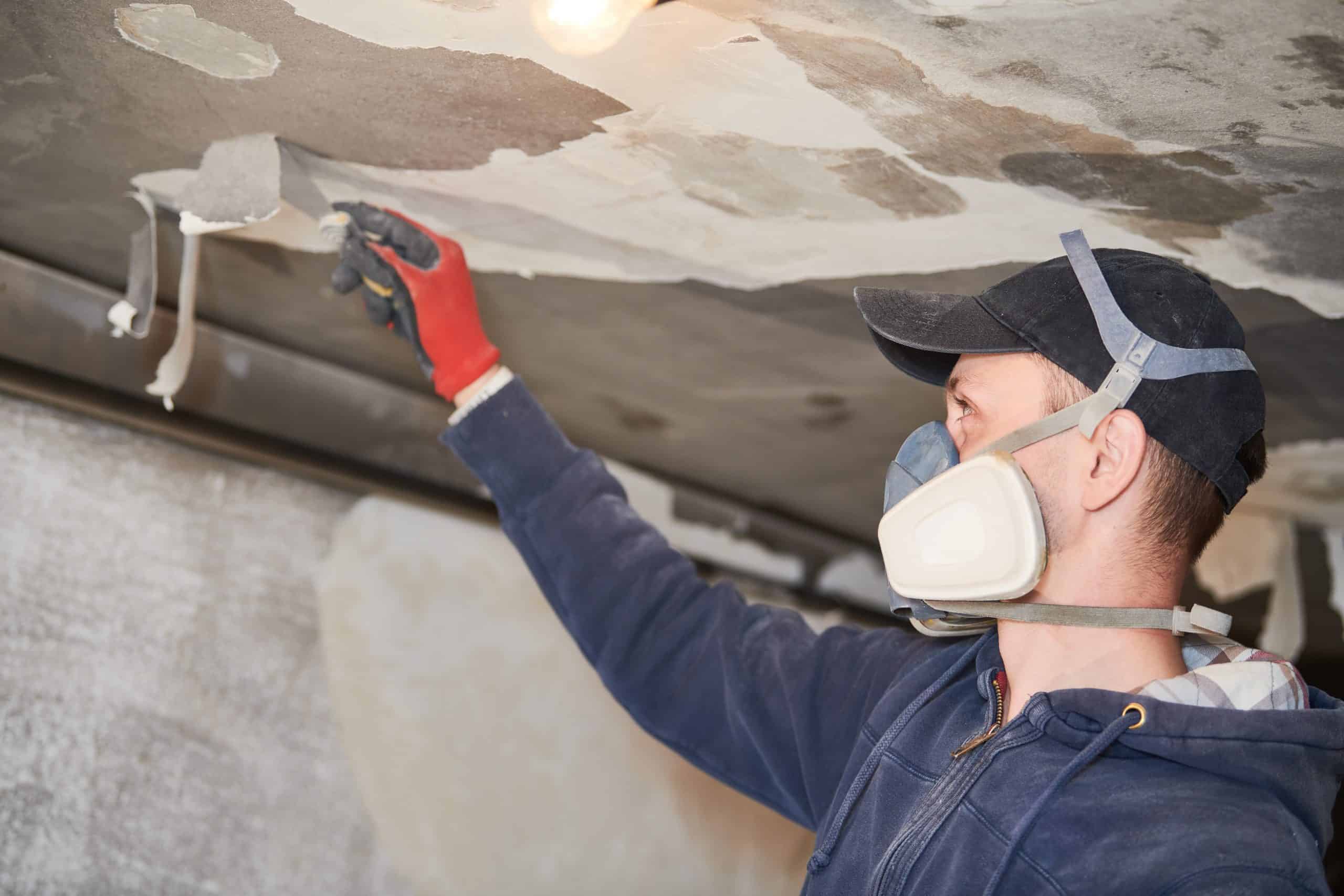 A person wearing a cap, mask, and gloves is applying plaster to a ceiling with a trowel. The scene is in a dimly lit room with visible patches of unpainted areas on the ceiling.