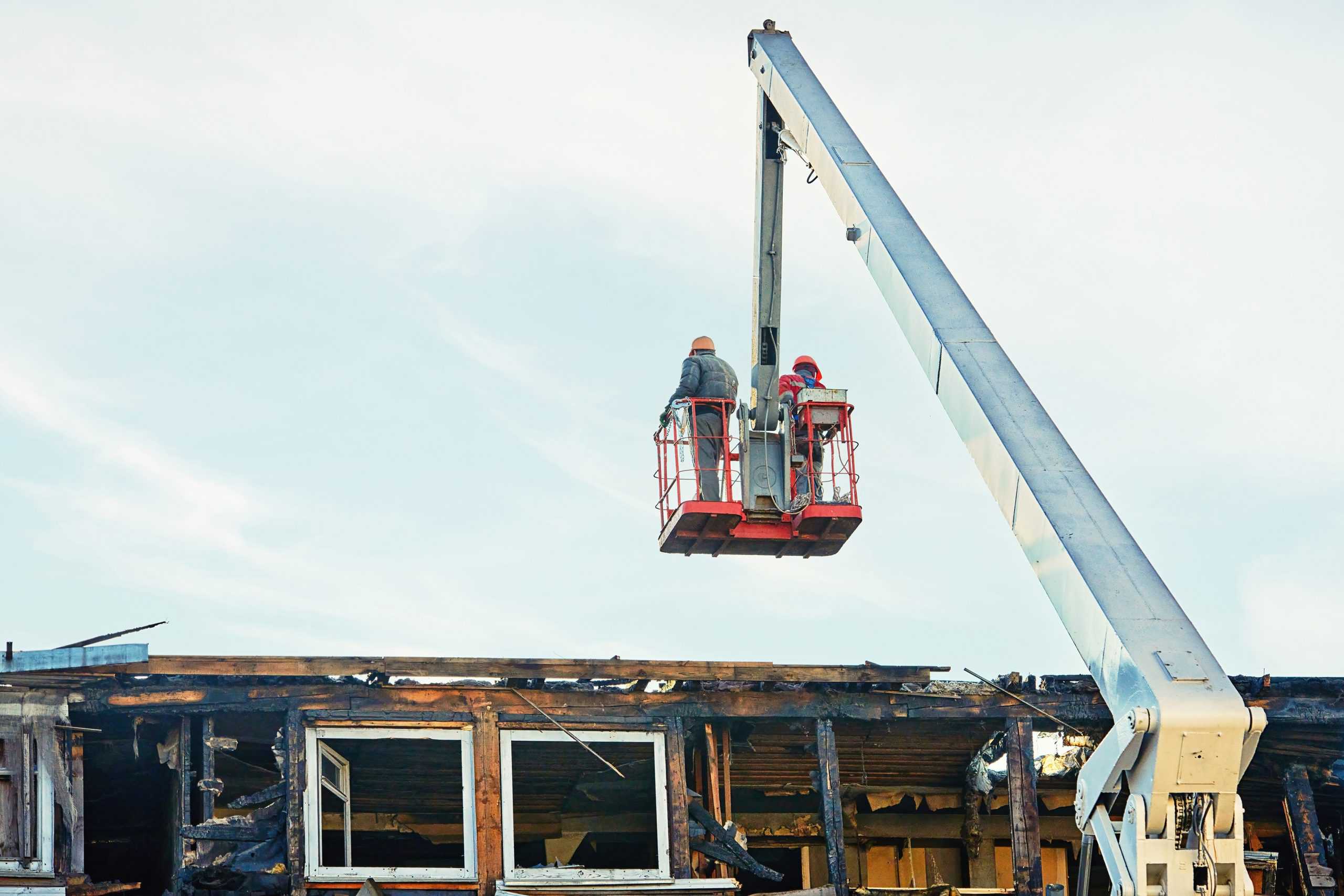 Two construction workers in a crane basket inspect the damaged upper section of a building. The structure shows signs of fire damage, with charred walls and broken windows. A clear sky is in the background.